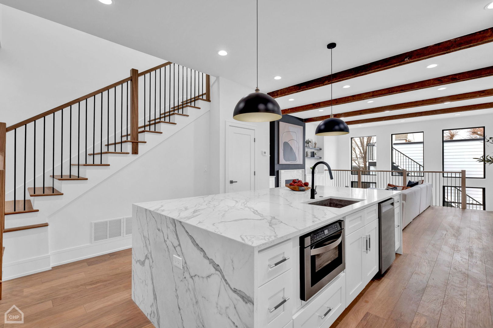 Open-plan kitchen with a large marble island, exposed wood beams, and a wood-and-metal staircase.
