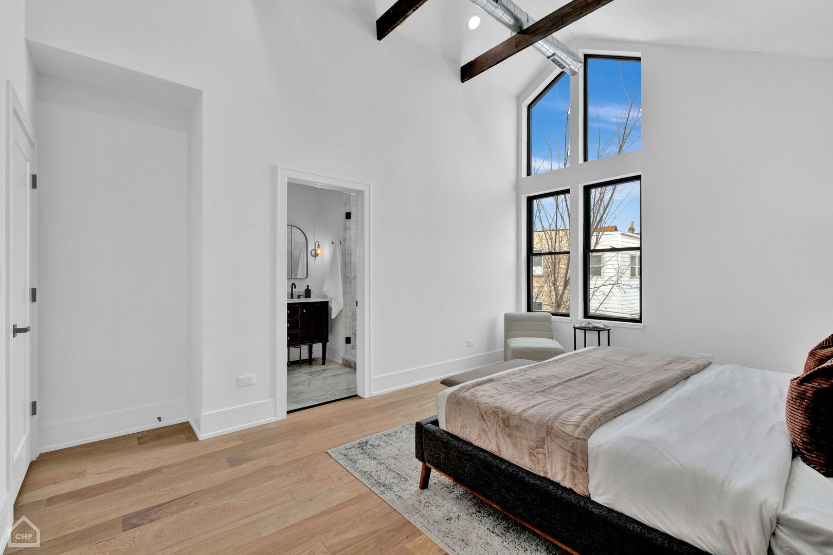 Bedroom with vaulted ceiling, exposed beams, tall windows, and an open doorway to the bathroom.