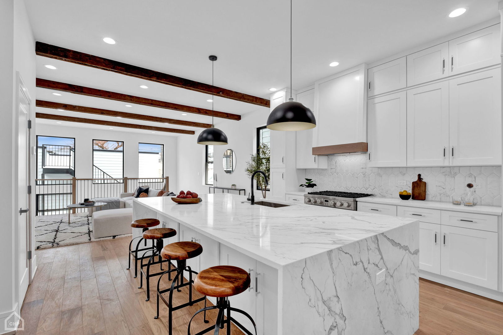 Bright open kitchen with a marble waterfall island, wood-topped stools, white cabinetry, and exposed ceiling beams.