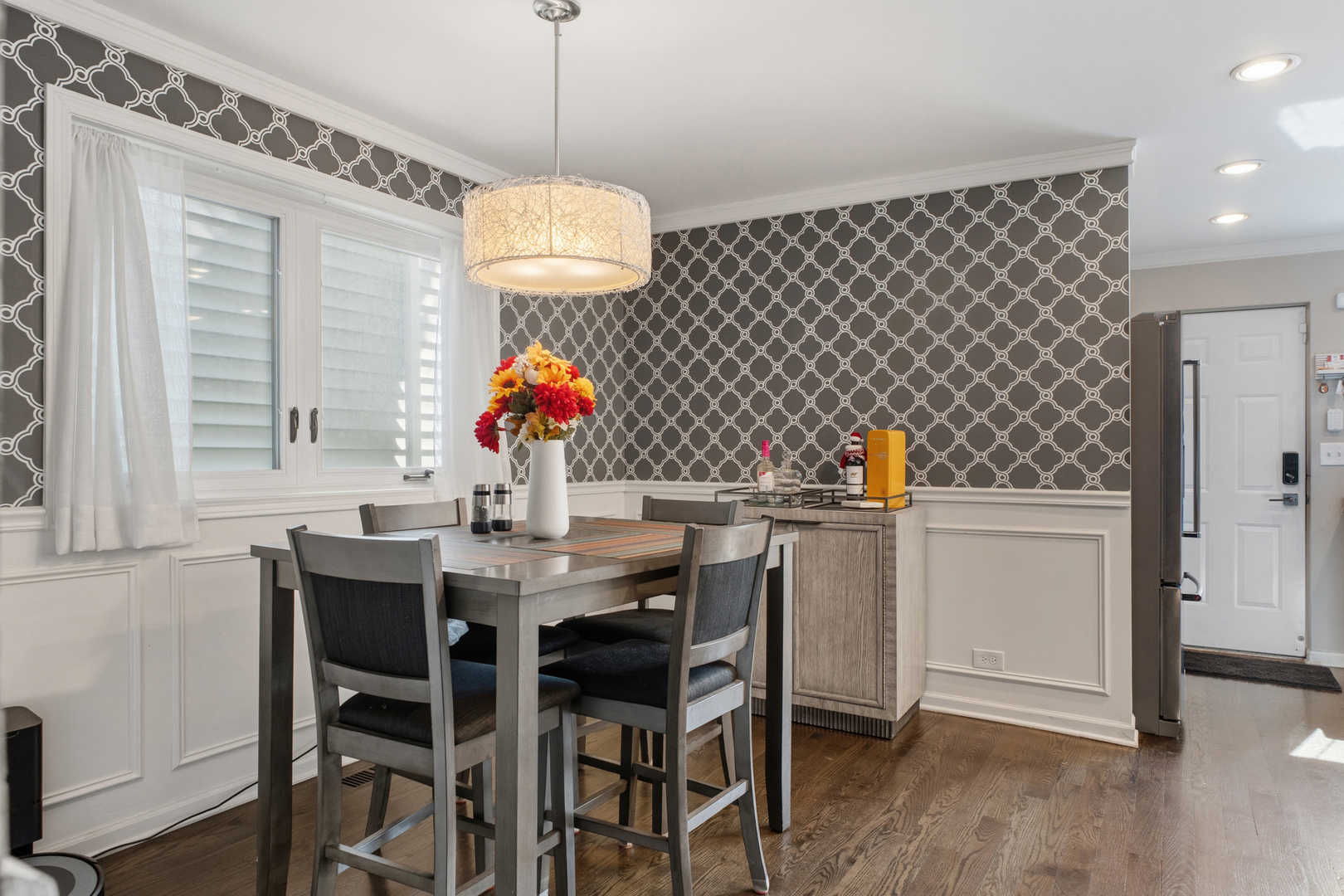 Dining area with patterned wallpaper, a gray table and chairs, curtained windows, and hardwood floors.
