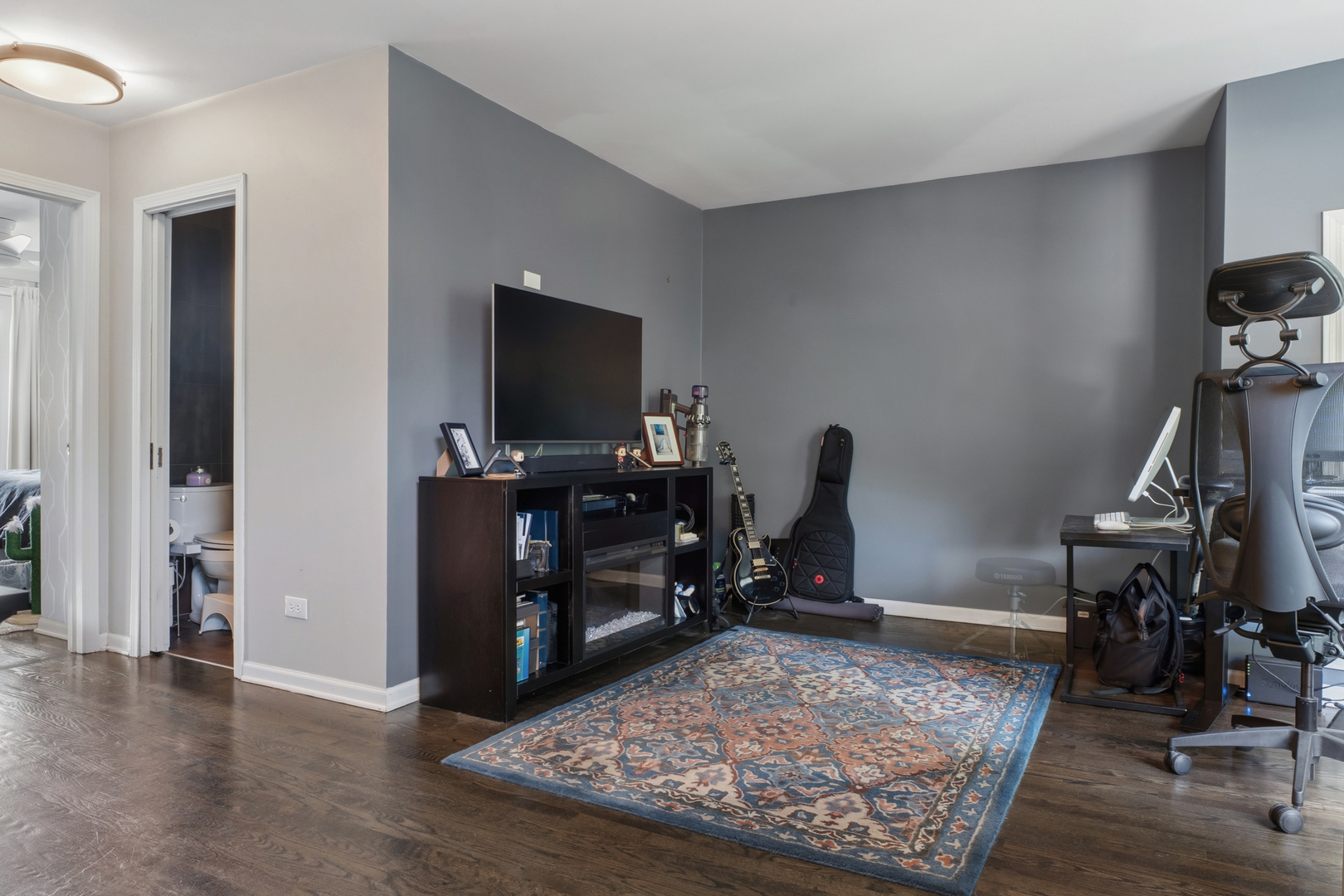 Furnished home office with a guitar, TV, desk, and patterned blue rug.