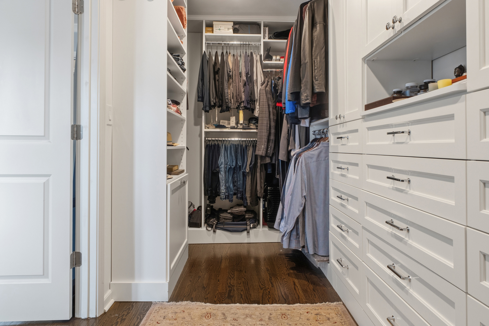 Walk-in closet with white built-in drawers, shelves, hanging clothes, and dark hardwood flooring.