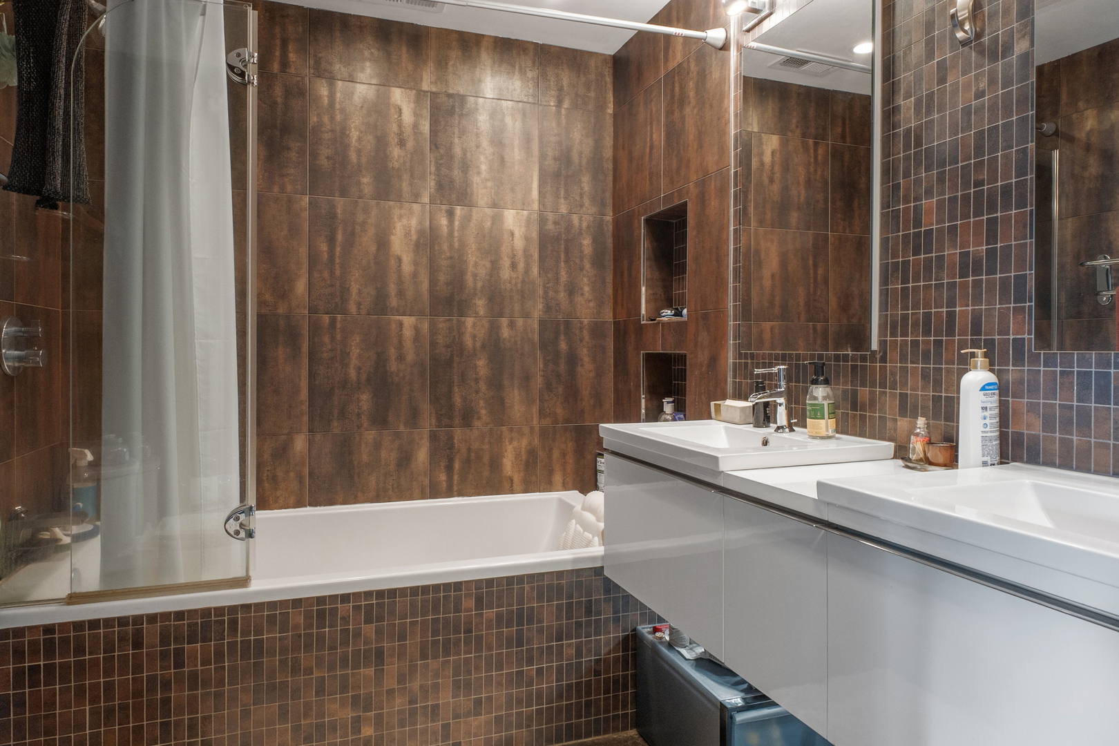 Bathroom with a glass-enclosed tub, brown tile, and a double-sink white vanity.