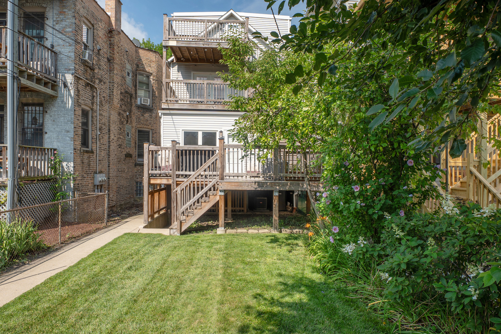 Home’s rear decks and stairs overlook a grassy backyard with mature trees and flowering garden beds.
