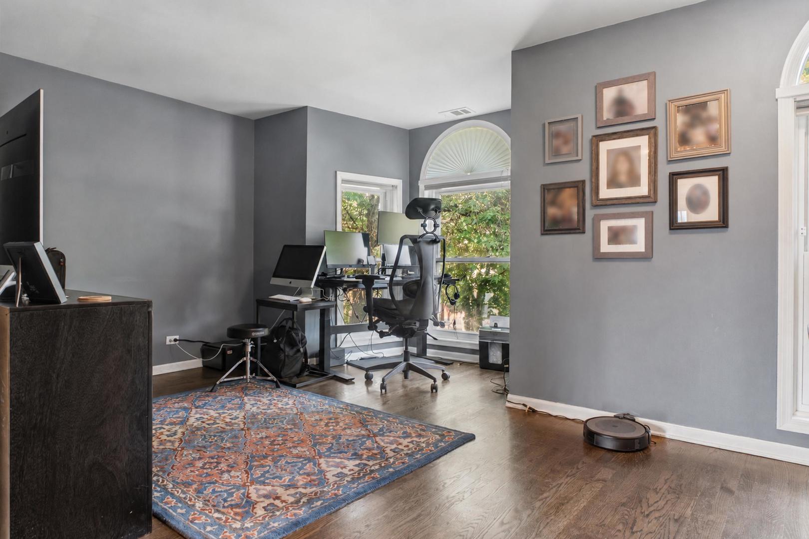 Home office with computer desks, arched windows, gray walls, framed artwork, and a patterned rug.
