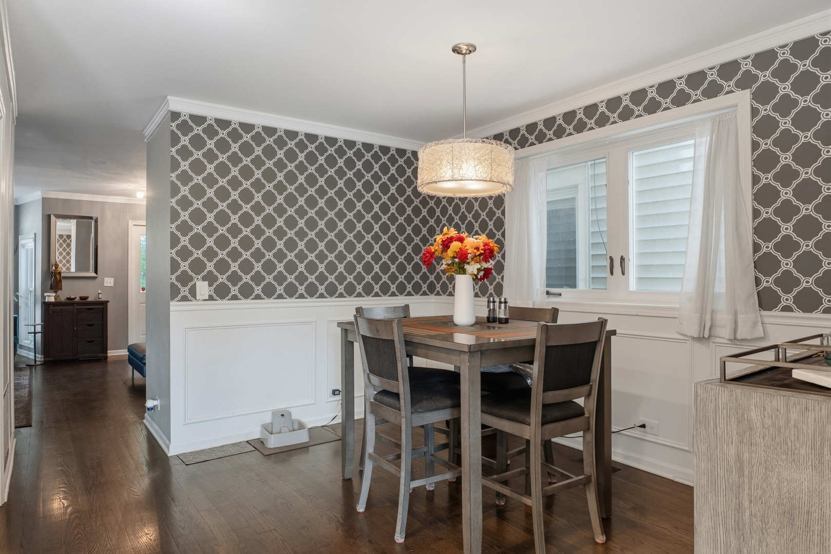 Dining room with patterned wallpaper, white wainscoting, and a dark wood table beneath a pendant light.