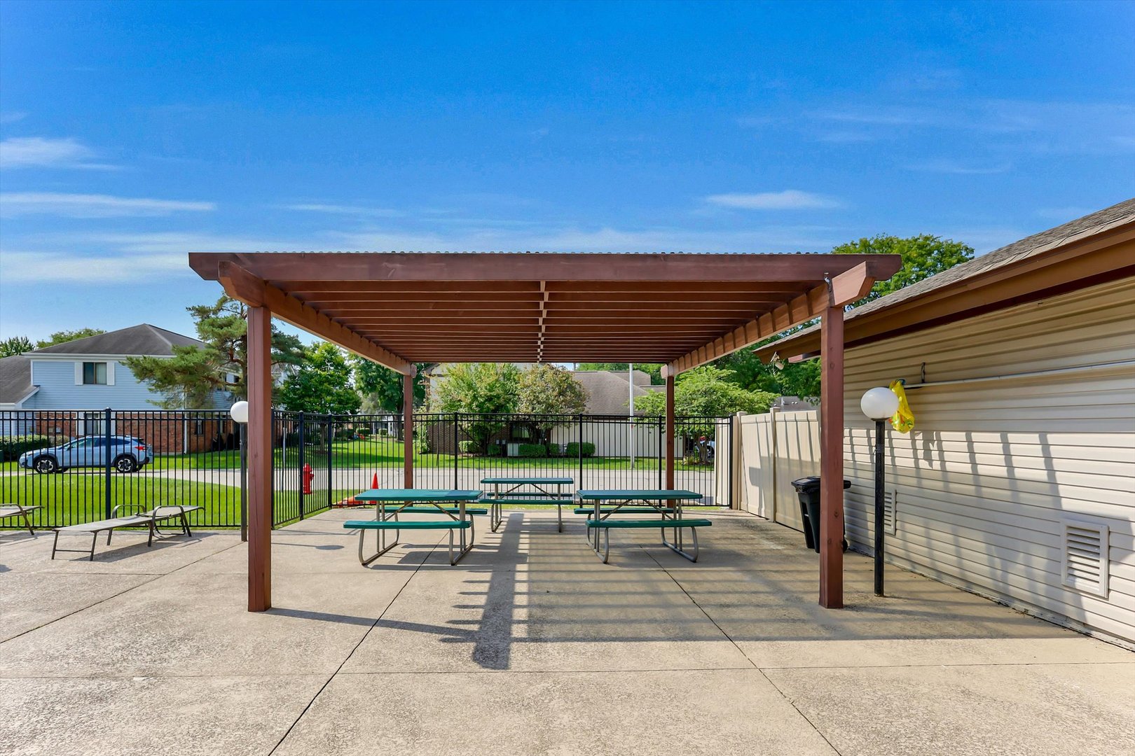 Covered patio with a wooden pergola and picnic tables beside a fenced lawn.