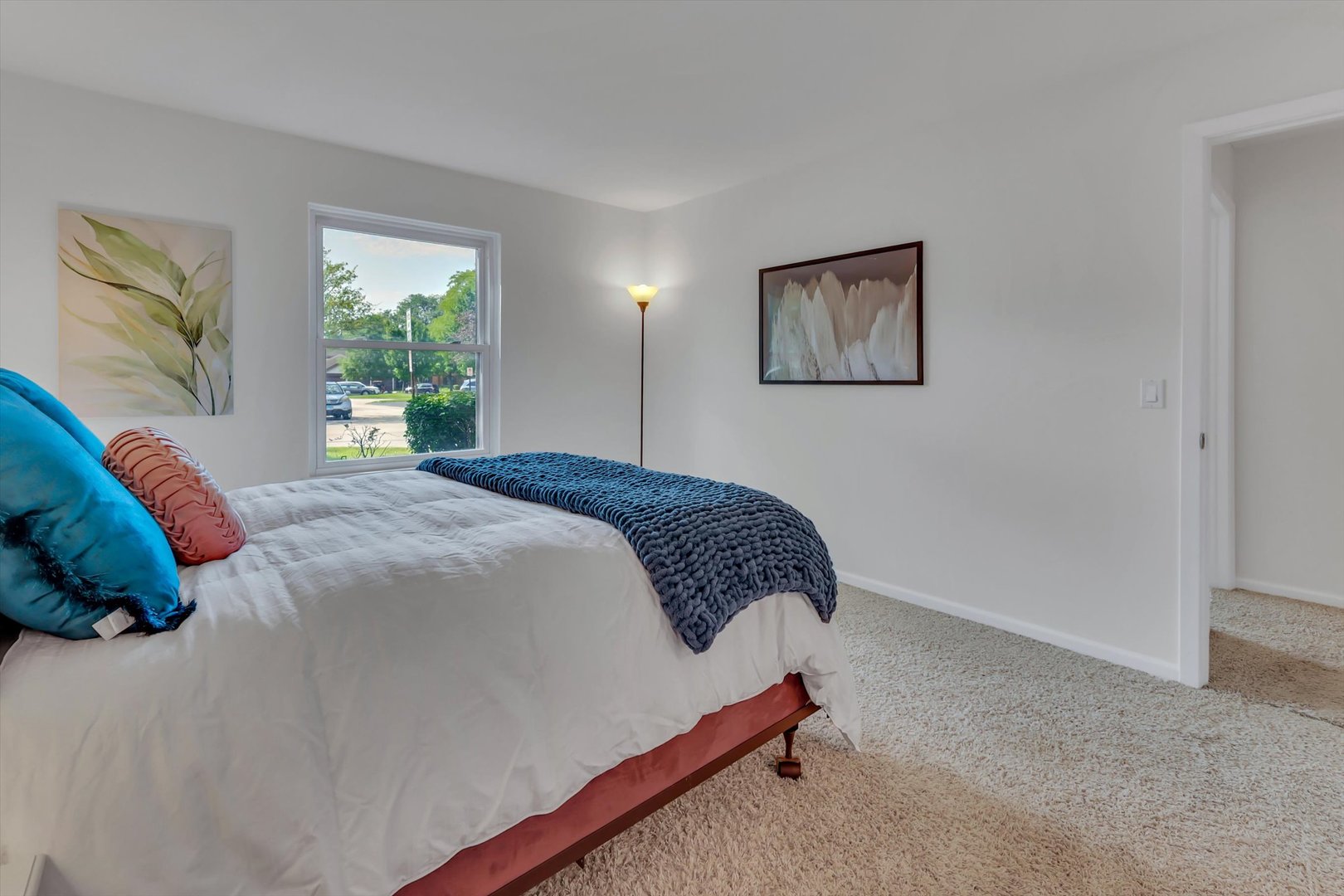 Staged bedroom with a white bed, blue throw, plush carpet, and a window overlooking trees.