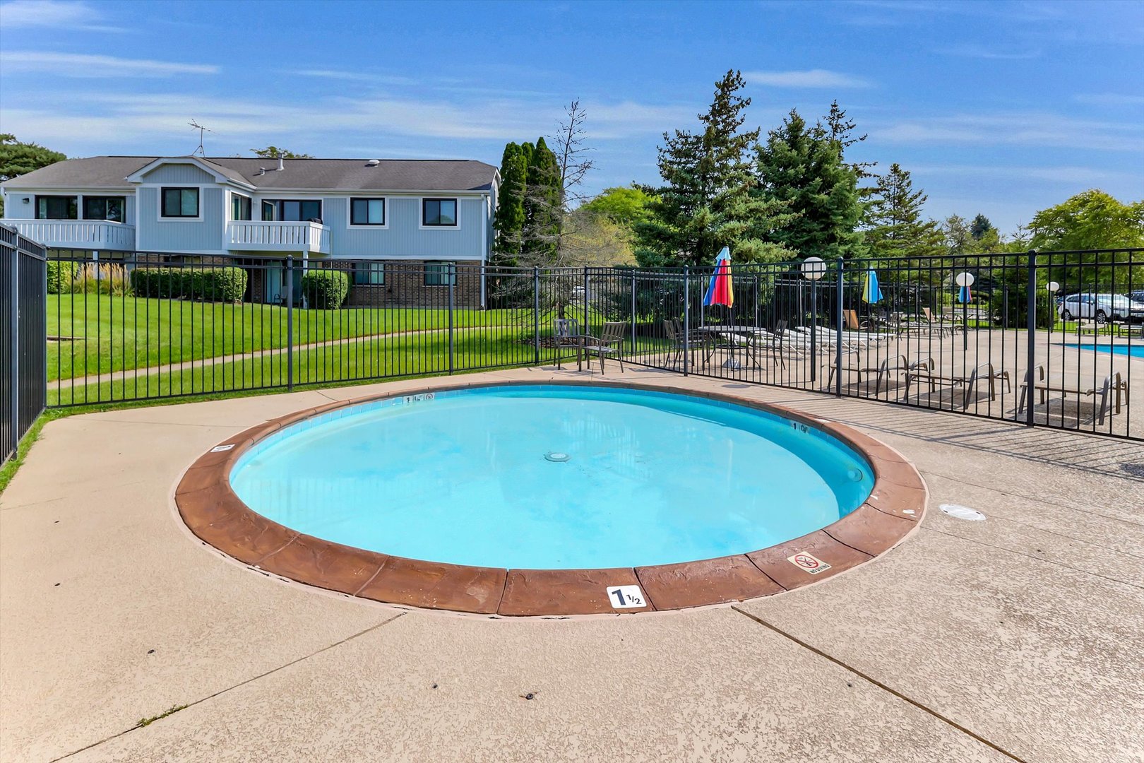 Outdoor community pool with a shallow rounded section, lounge chairs, and umbrellas beside a residential building.
