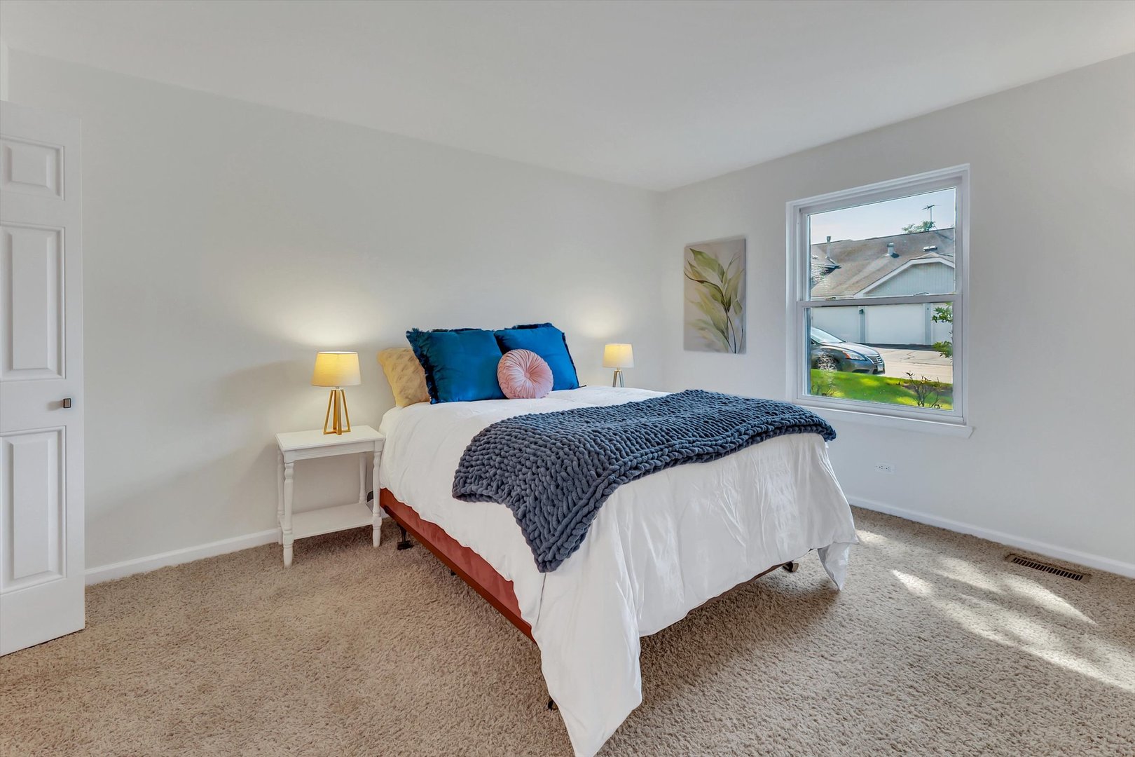 Staged bedroom with a white bed, blue pillows, a chunky gray throw, and a window overlooking neighboring homes.