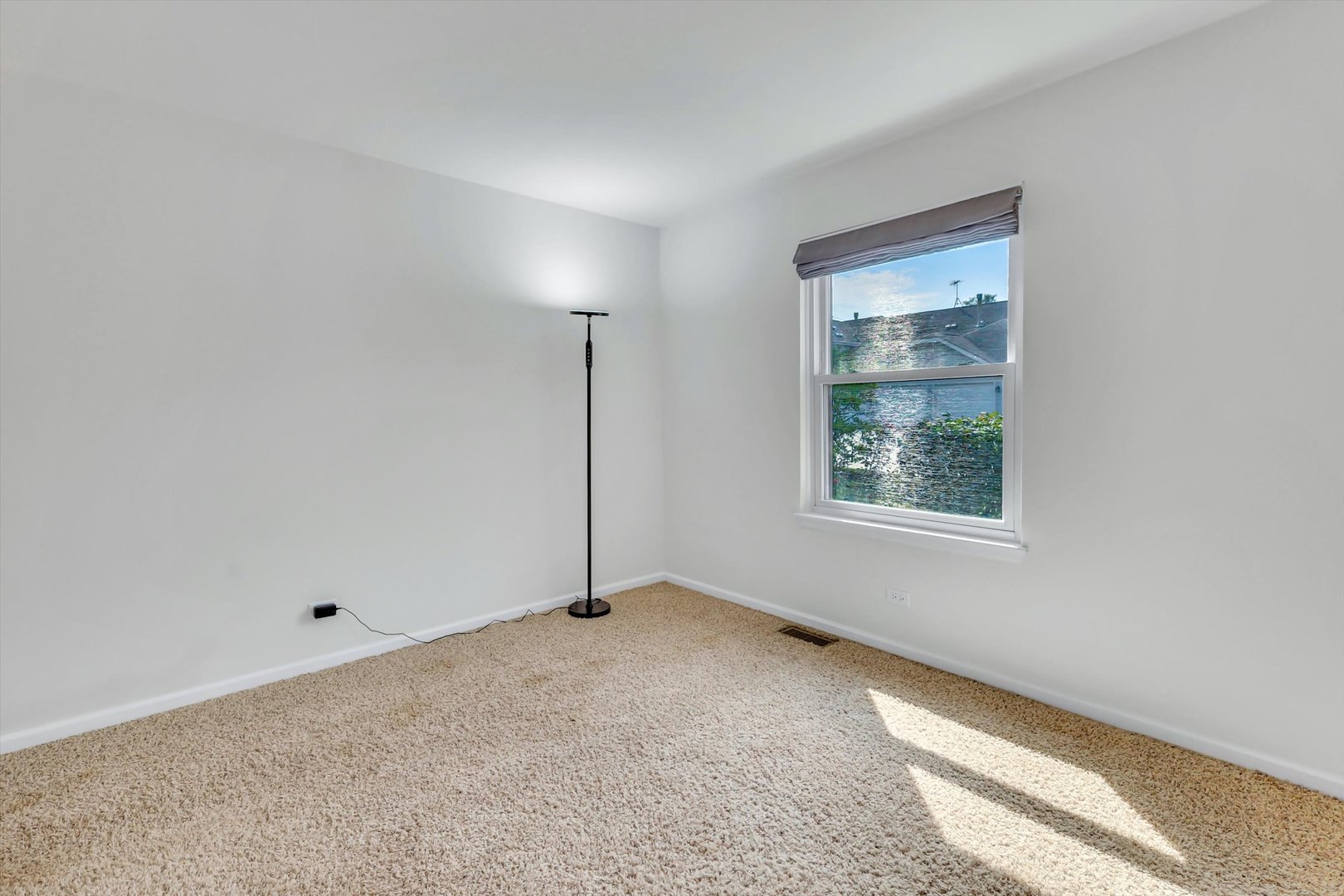 Empty carpeted room with white walls, a floor lamp, and sunlight through one window.