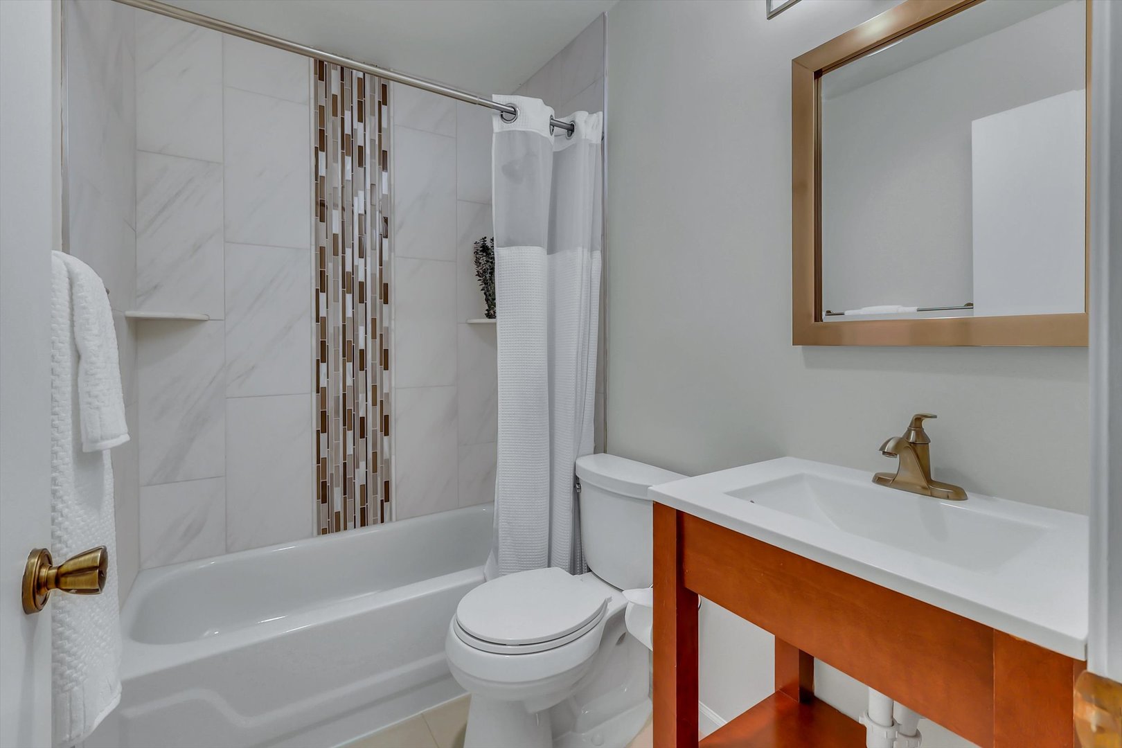 Bathroom with a tiled tub and shower, mosaic accent strip, and wood vanity with bronze fixtures.