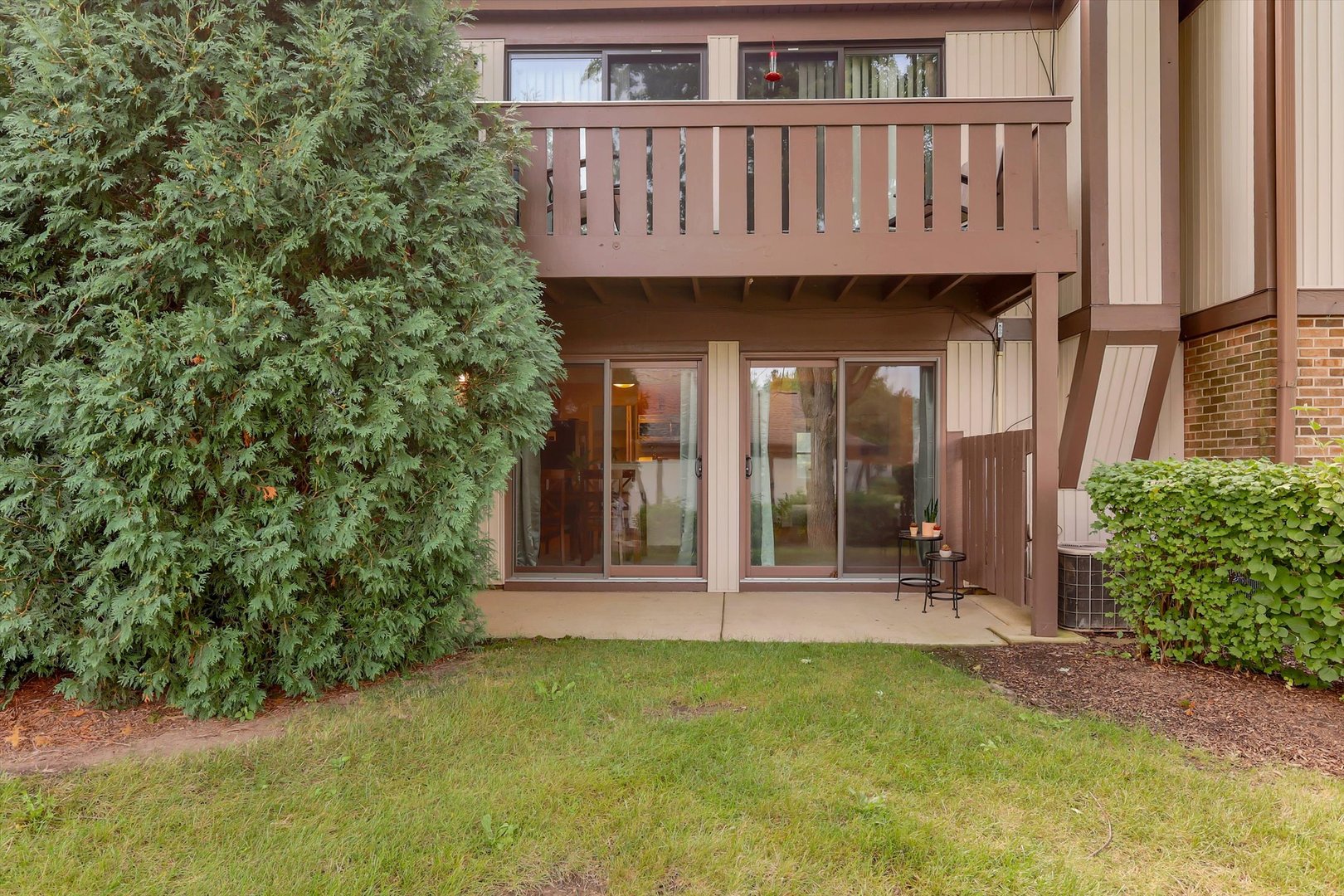 Condominium rear patio with sliding glass doors beneath an upper-level balcony, partly screened by an evergreen shrub.