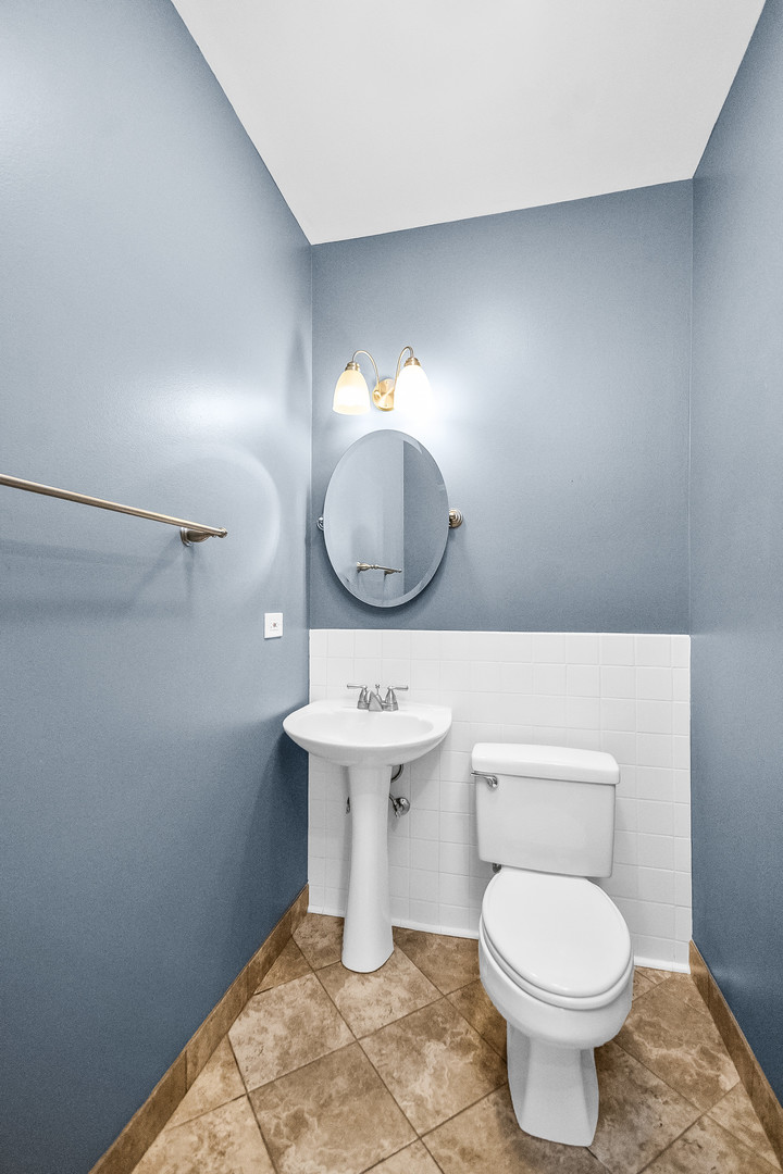 Bathroom with blue-gray walls, white tile wainscoting, pedestal sink, oval mirror, and brown patterned floor.