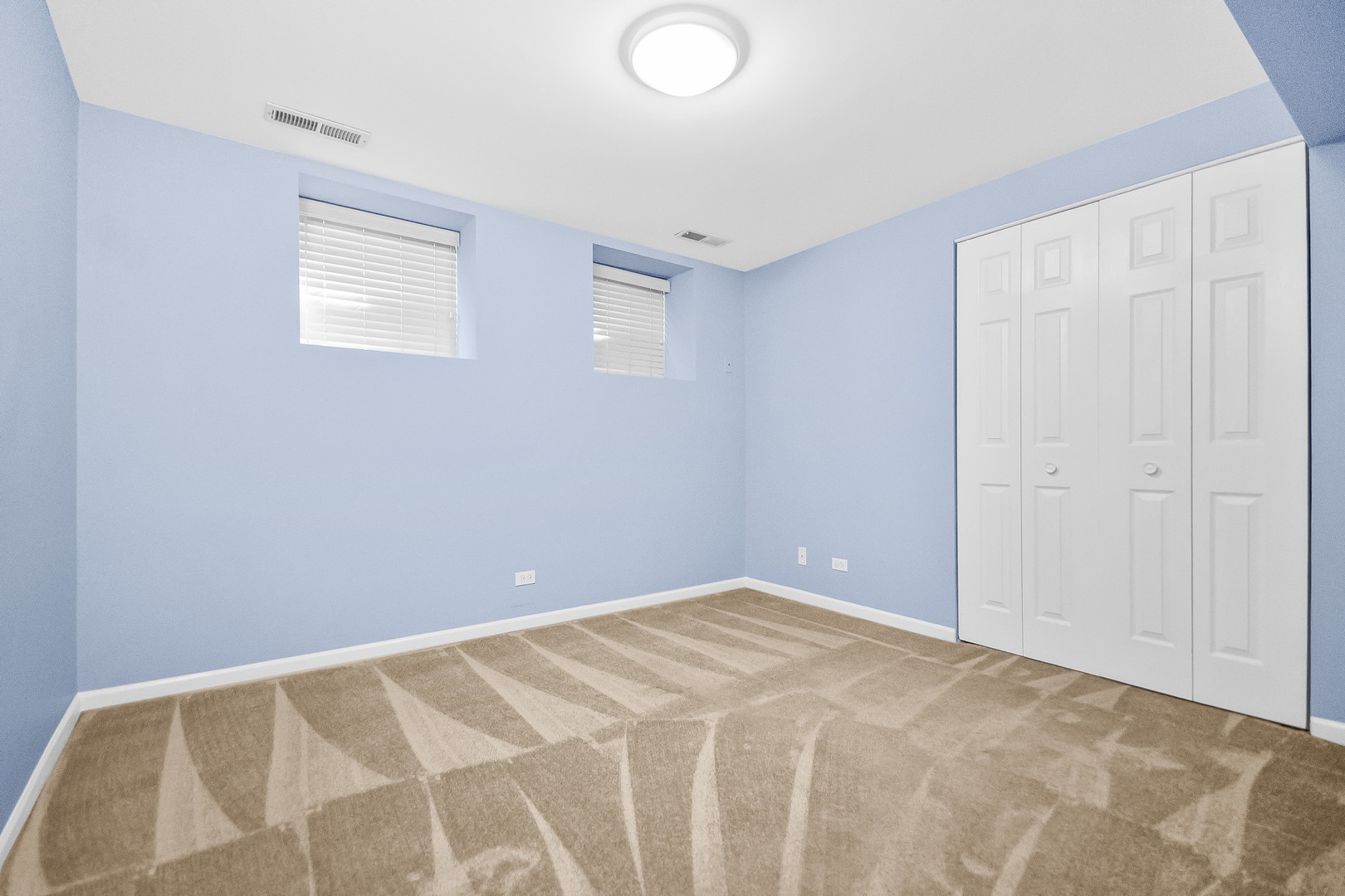 Empty bedroom with pale blue walls, beige carpet, two high windows, and white closets.
