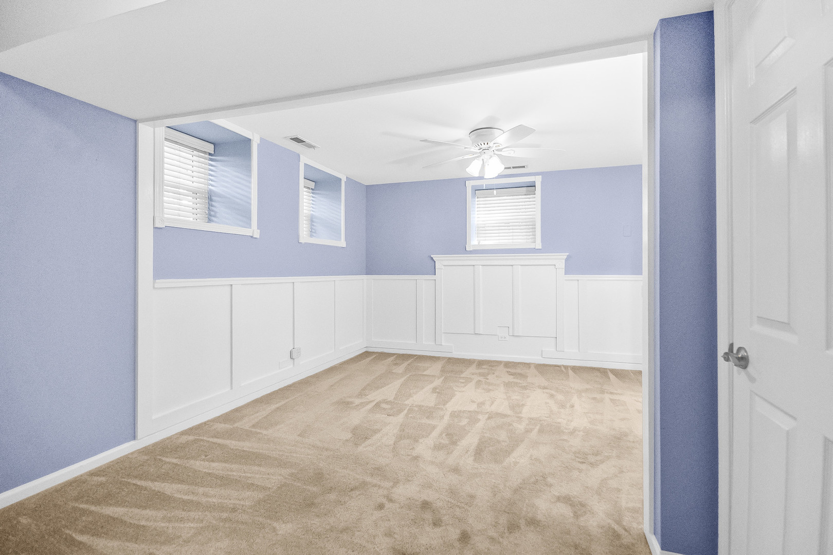 Empty carpeted room with blue walls, white wainscoting, high windows, and a ceiling fan.