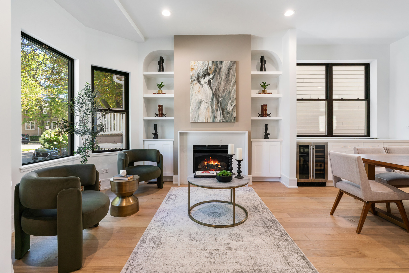 Furnished living room with a lit fireplace, built-in shelves, and large black-framed windows.