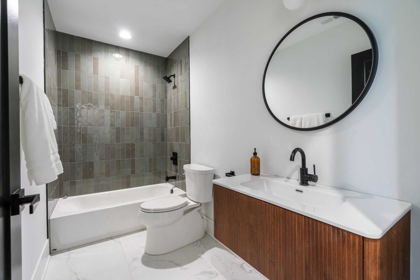 Modern bathroom with a gray-green tiled tub and shower, wood vanity, and round mirror.