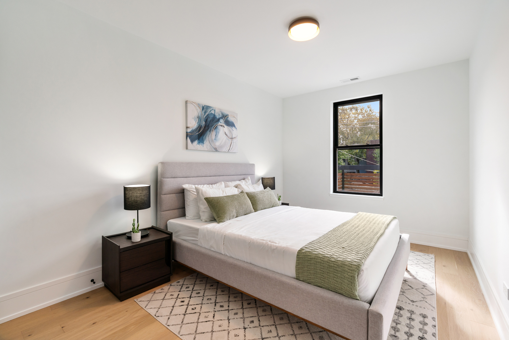 Staged bedroom with a gray upholstered bed, green accents, and a black-framed window.