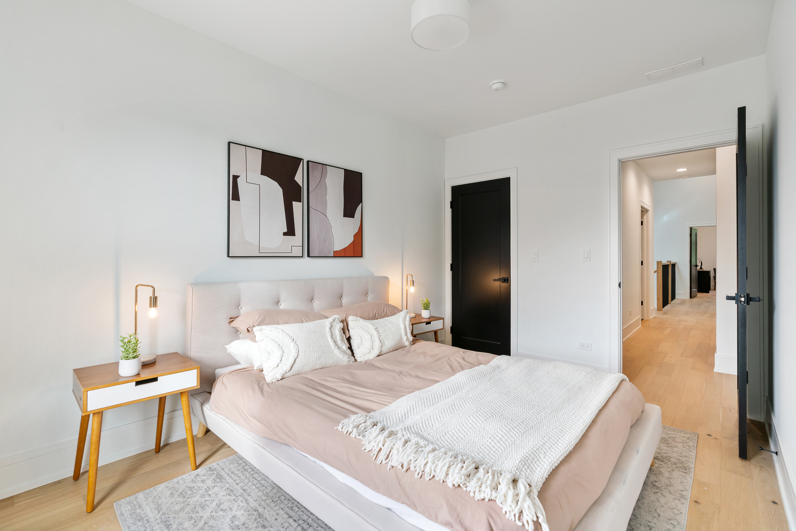 Staged bedroom with a blush-covered bed, abstract wall art, and an open doorway to the hallway.