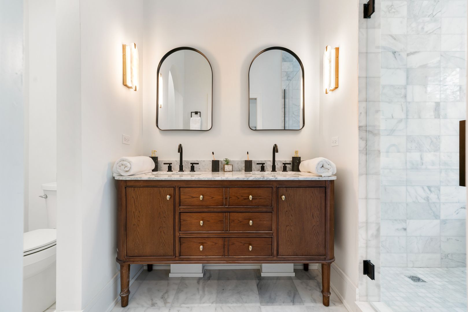 Bathroom with a dark wood double vanity, arched mirrors, and a marble-tiled glass shower.