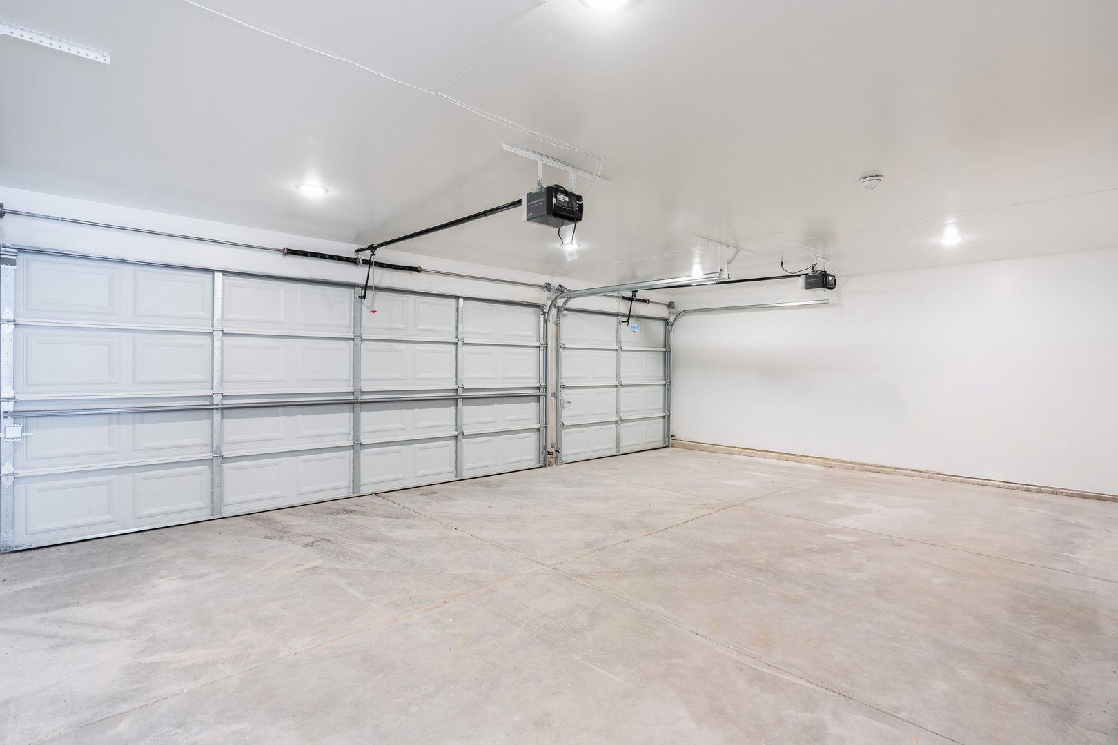 Empty garage with two white doors, ceiling-mounted openers, and a concrete floor.