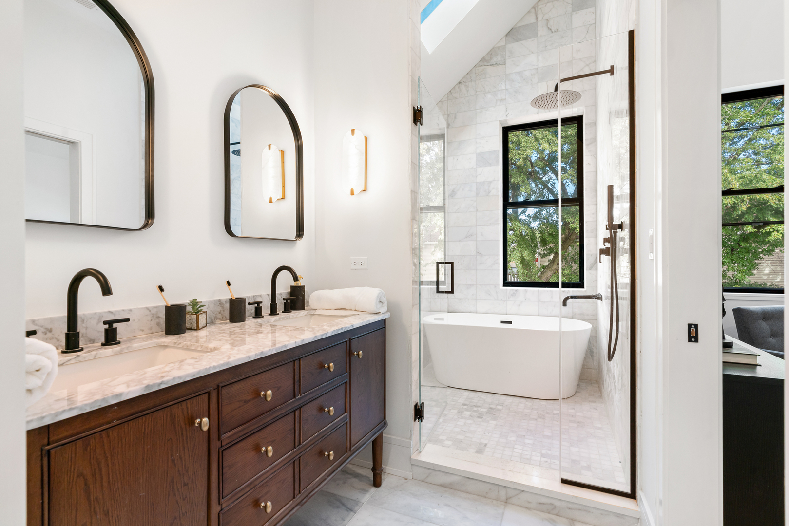 Primary bathroom with a dark wood double vanity, marble shower, and freestanding tub beside a large window.