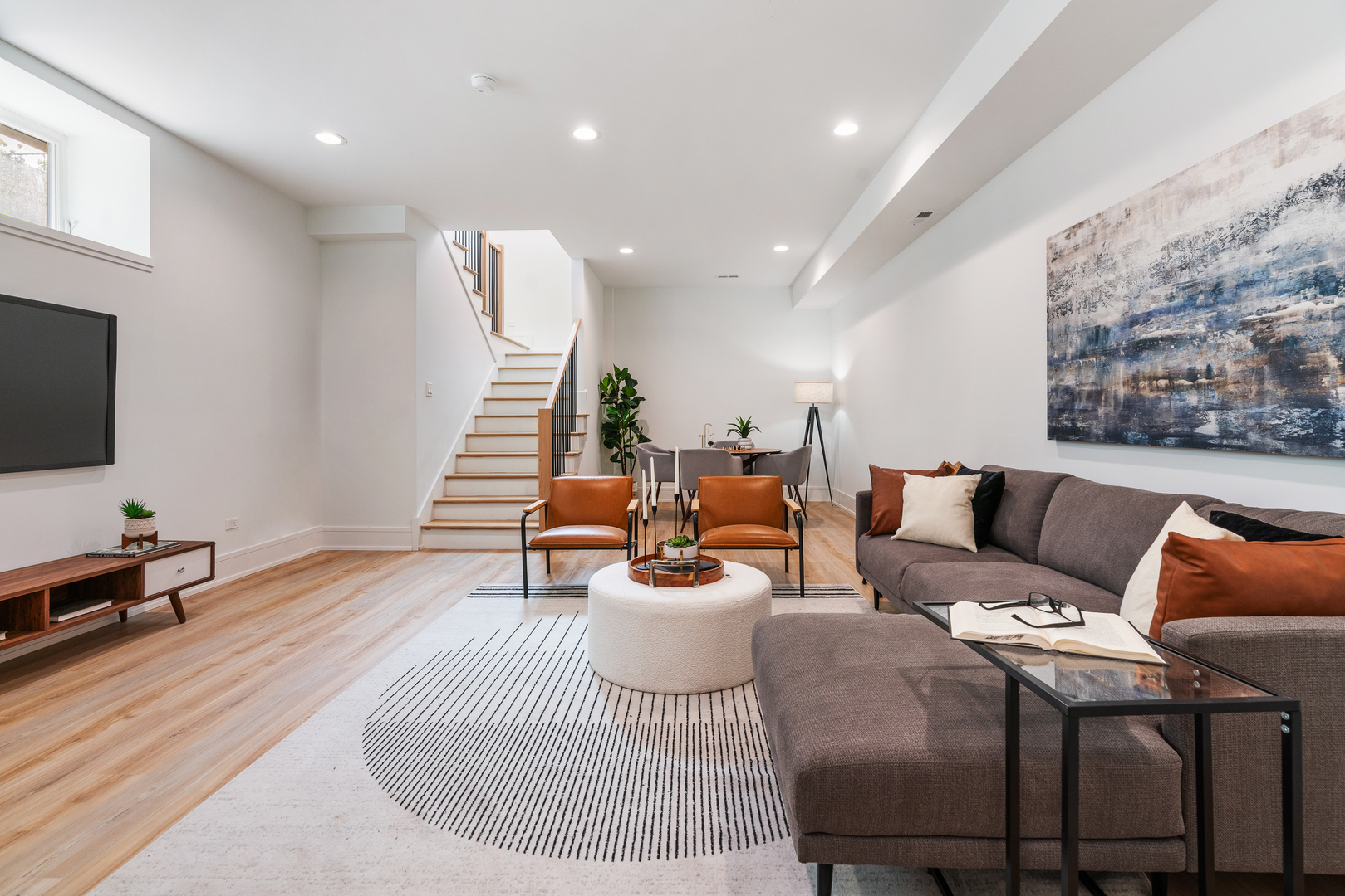 Staged living room with gray sectional, leather chairs, striped rug, and staircase beside dining area.