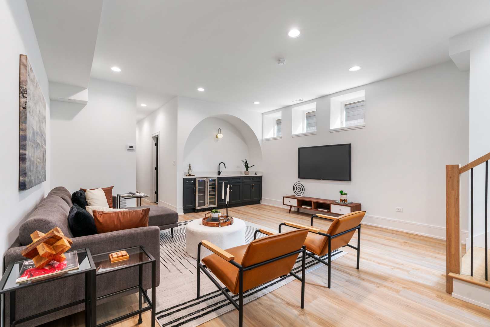 Staged living room with a dark-cabinet wet bar beneath an arch, leather chairs, and a wall-mounted TV.