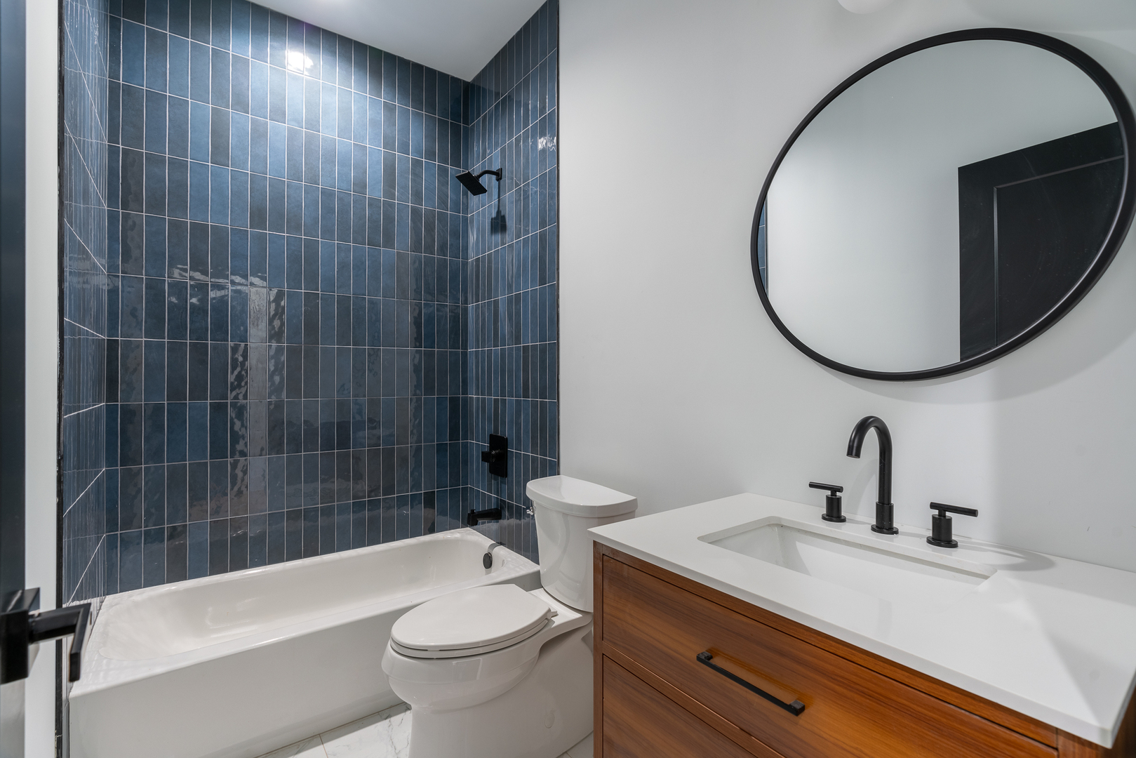 Bathroom with a dark blue tiled tub and shower, wood vanity, and large round mirror.