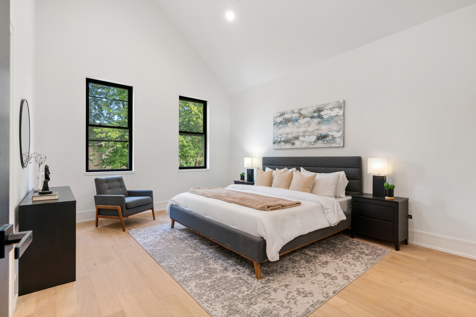 Staged bedroom with a vaulted ceiling, gray bed, pale wood floors, and two tall windows.