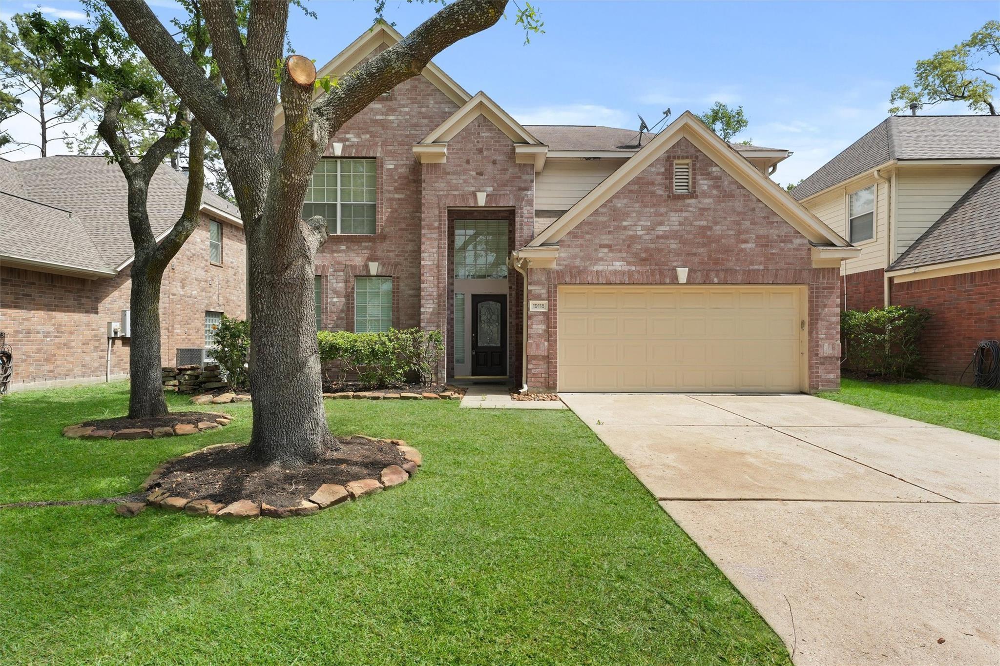 Two-story brick house with a tan garage door, broad driveway, and mature trees on the lawn.