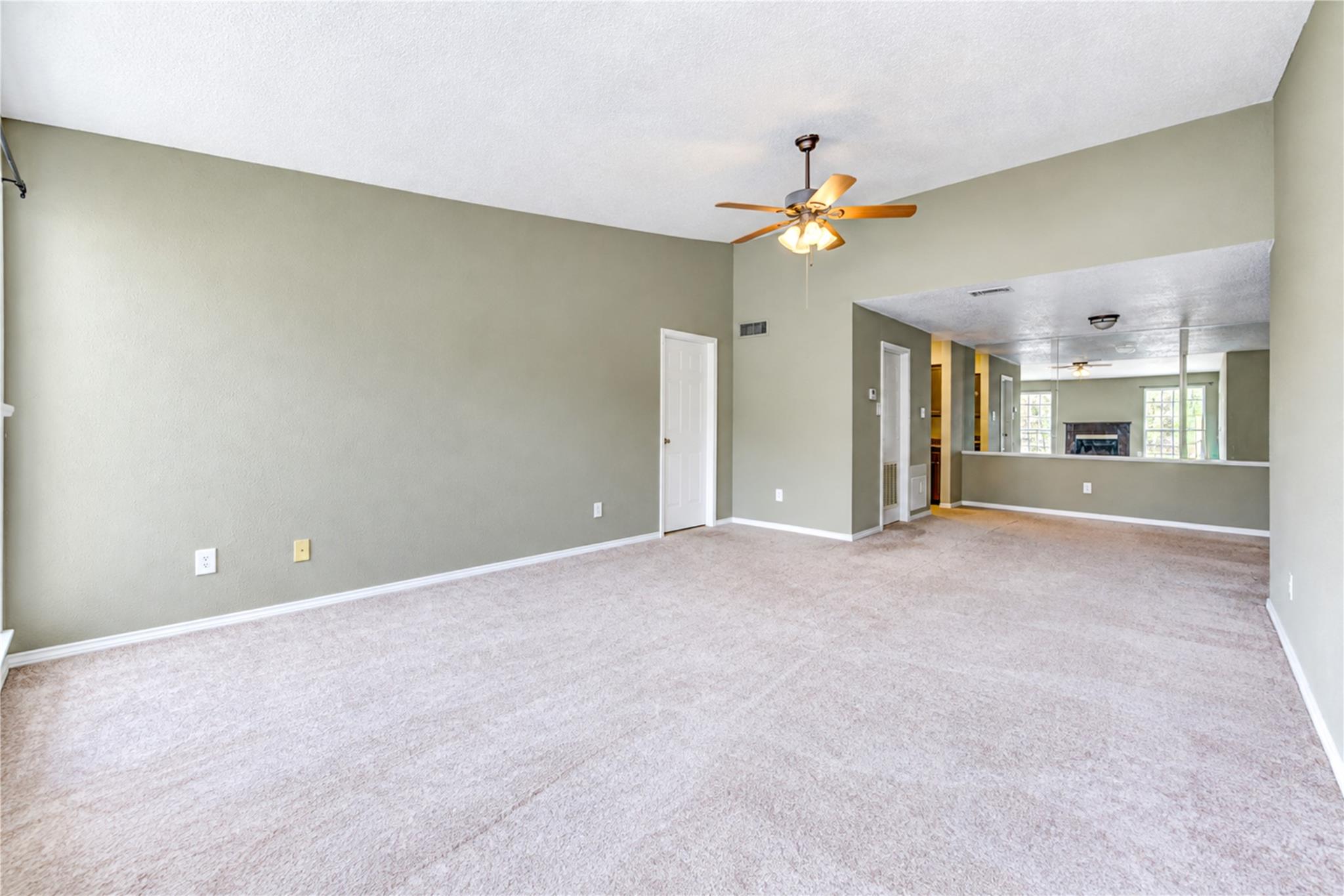 Empty living room with pale carpeting, a ceiling fan, and an adjoining room with a fireplace.