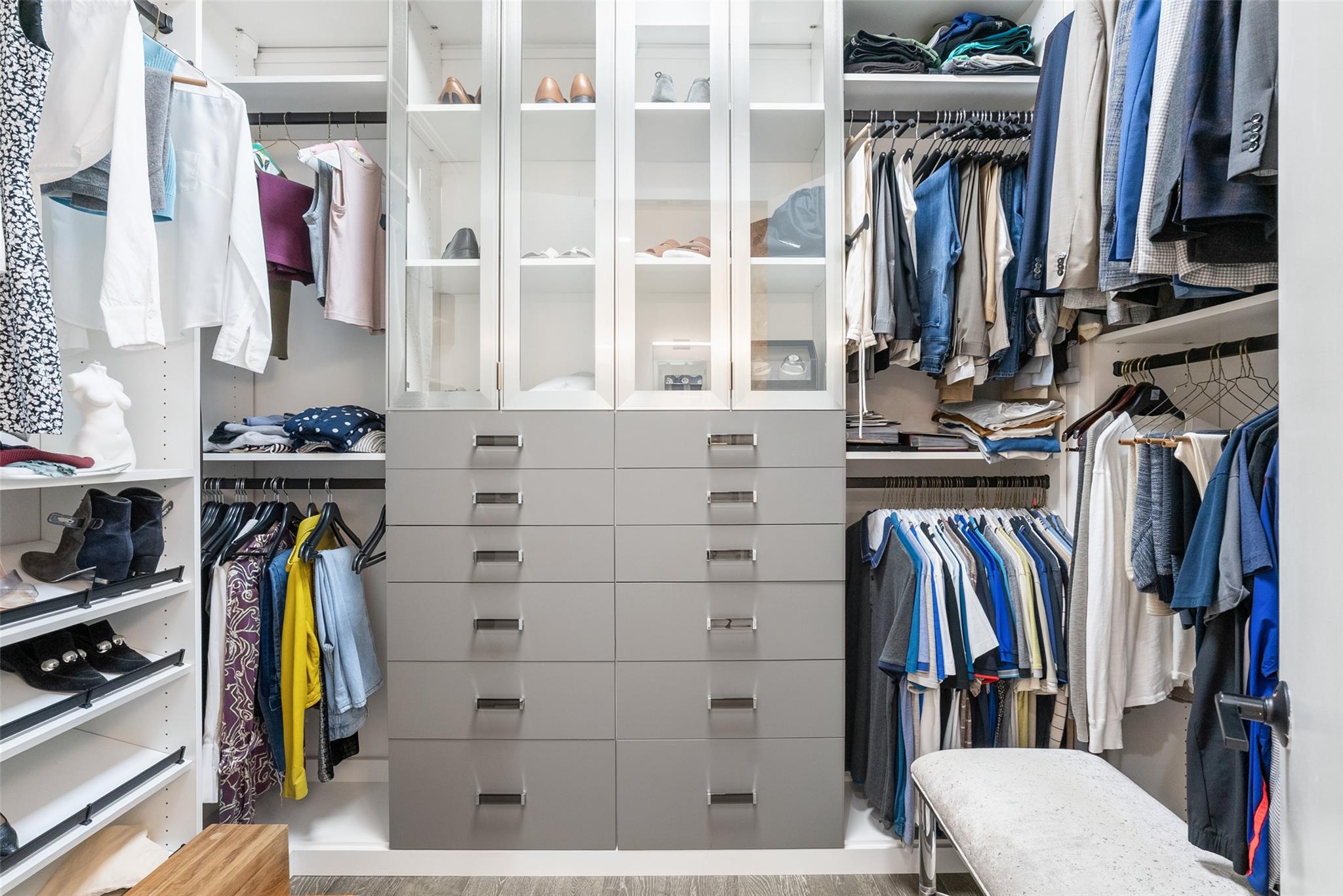 Walk-in closet with glass-front shoe storage, central drawers, hanging clothes, and a padded bench.