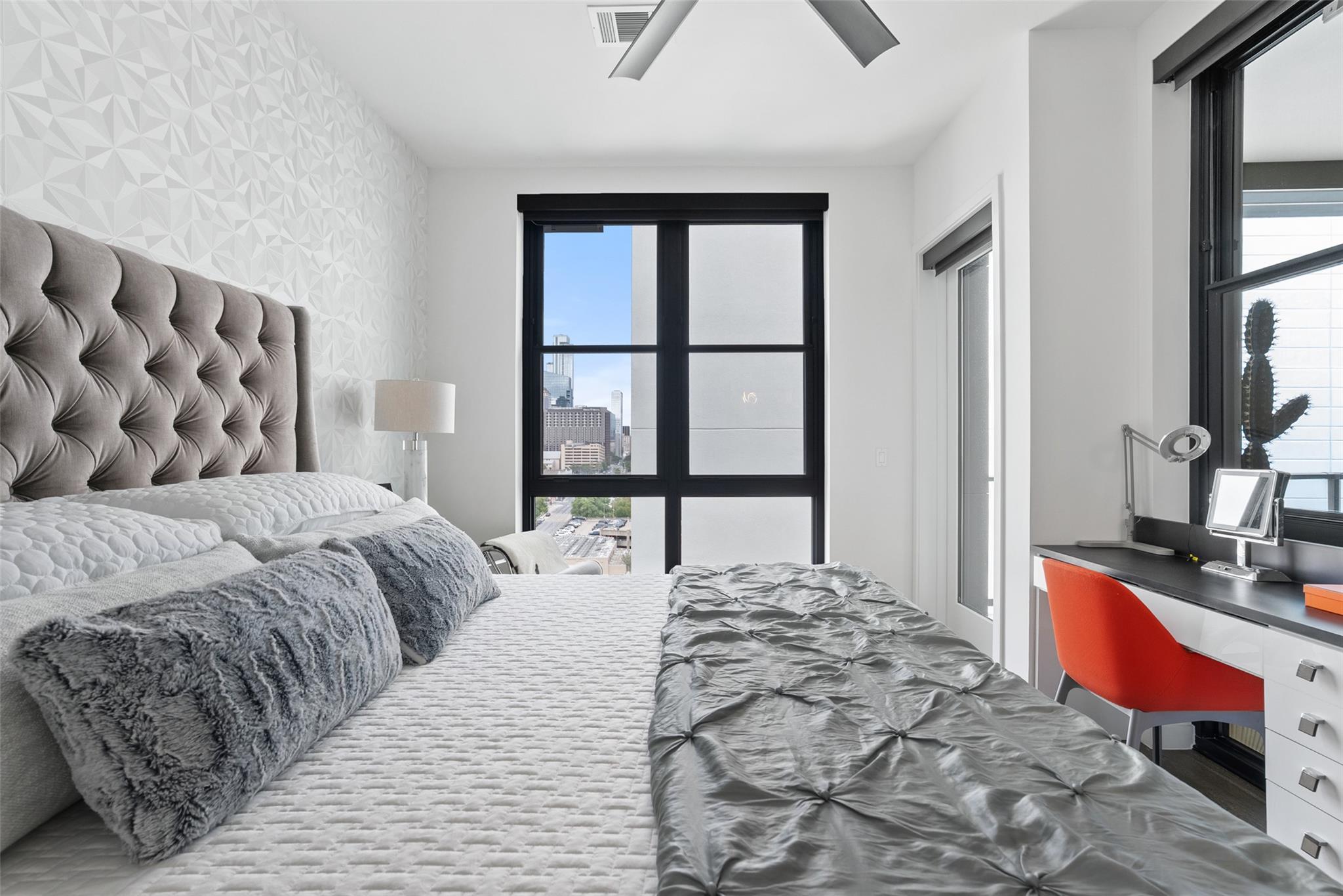 Modern bedroom with gray tufted headboard, red desk chair, and downtown views through black-framed windows.