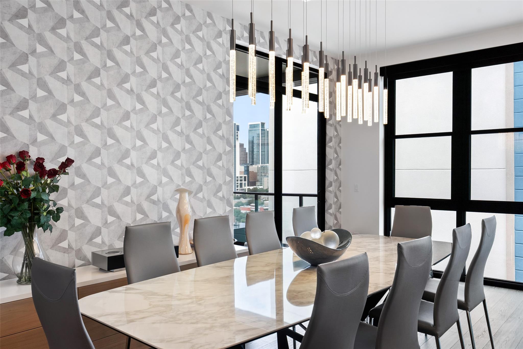Dining room with a marble table, geometric accent wall, pendant lights, and downtown views through tall windows.