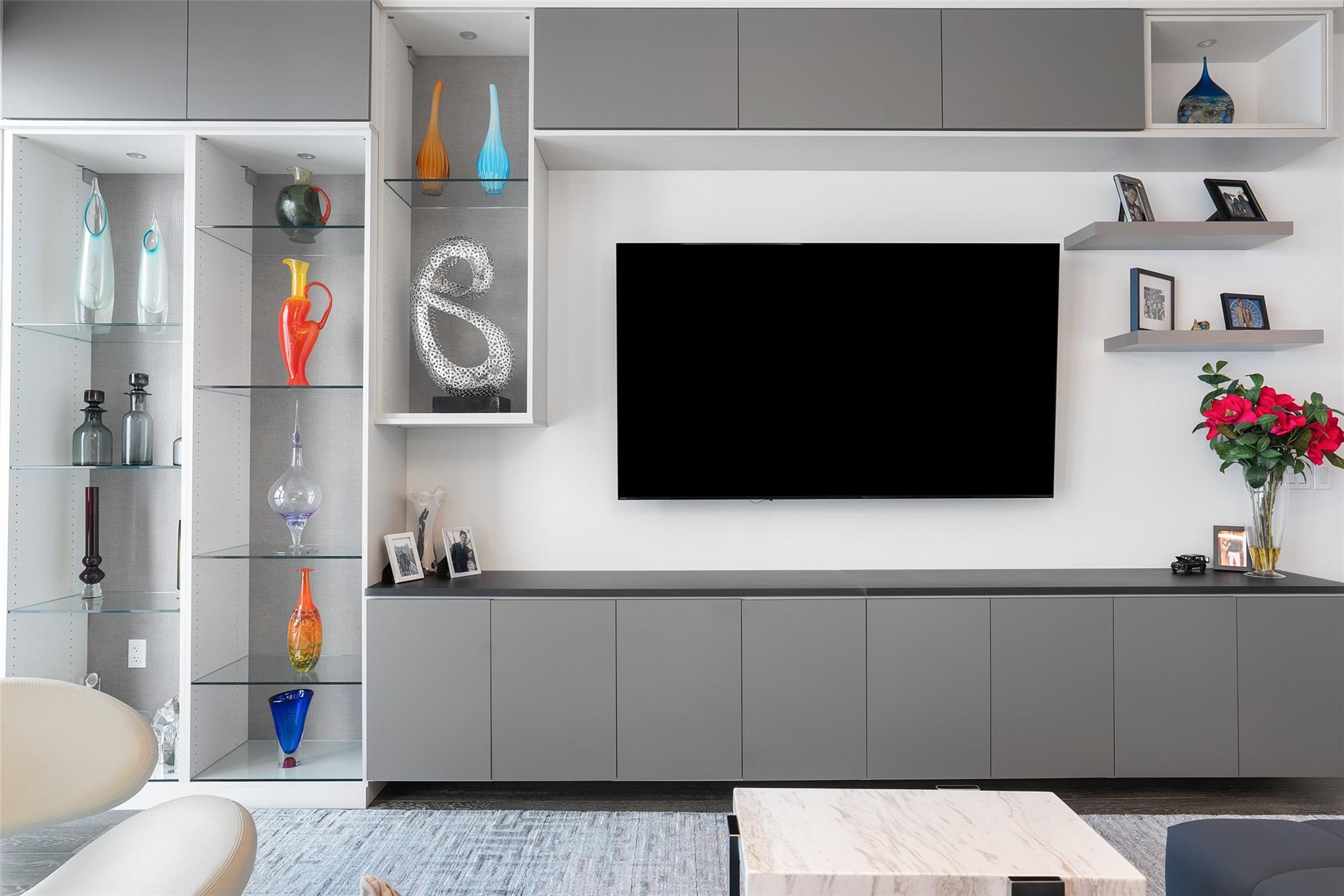 Living room with a large television, gray built-in cabinetry, and glass shelves displaying colorful vases.