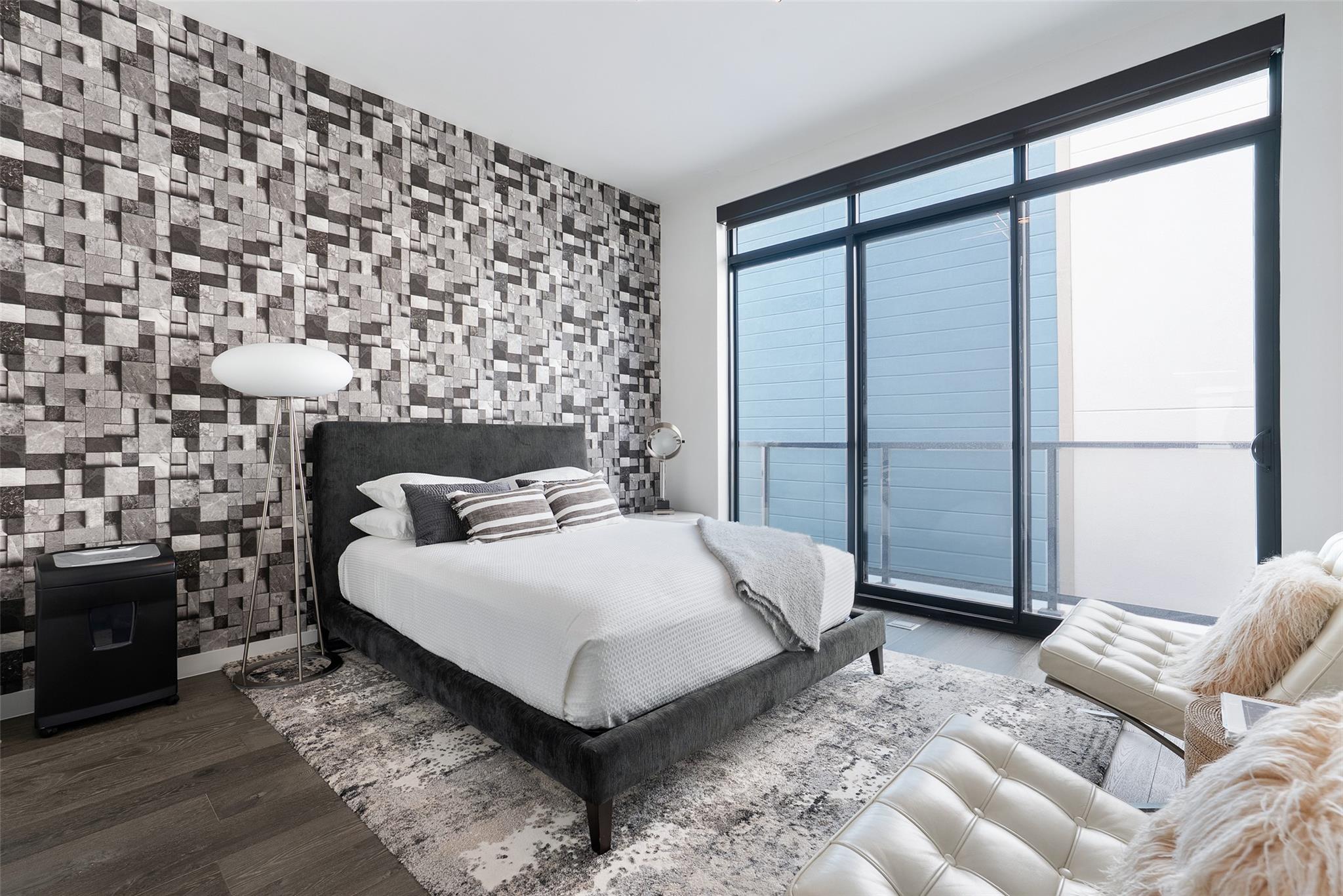 Modern bedroom with a charcoal bed, geometric accent wall, and floor-to-ceiling balcony windows.