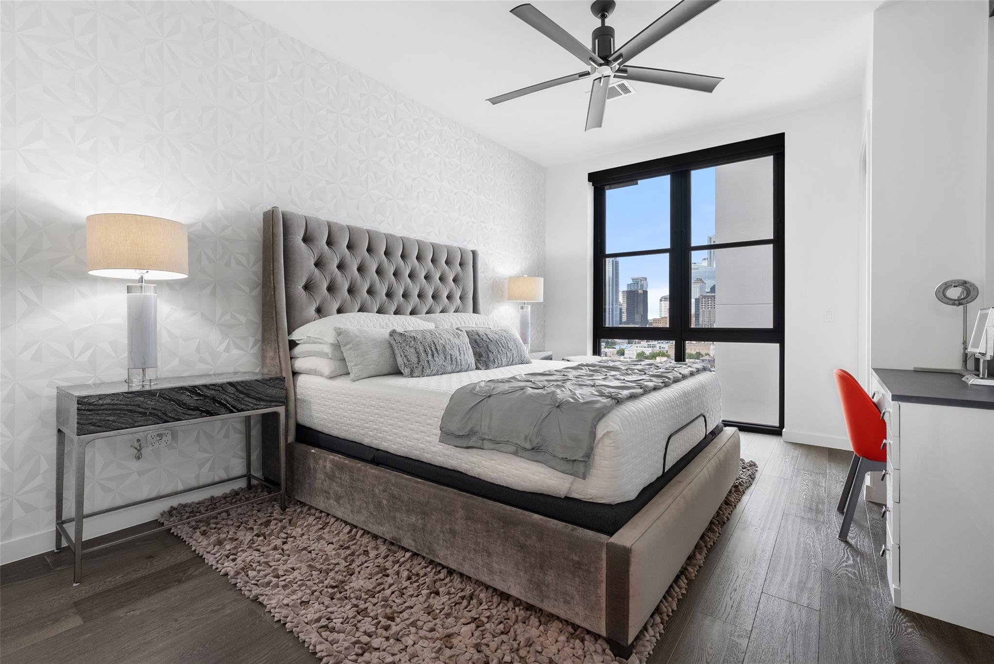 Modern bedroom with a tufted gray bed and downtown views through a large window.