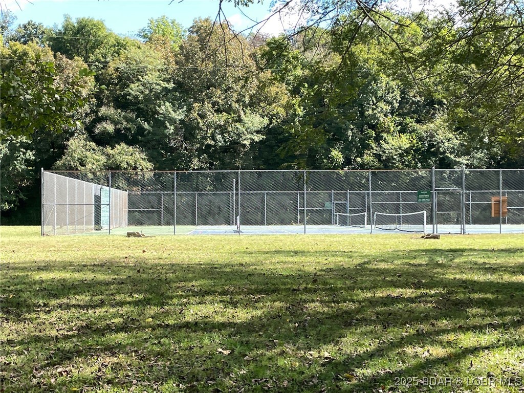 Fenced outdoor tennis court with a blue playing surface, grassy foreground, and wooded surroundings.