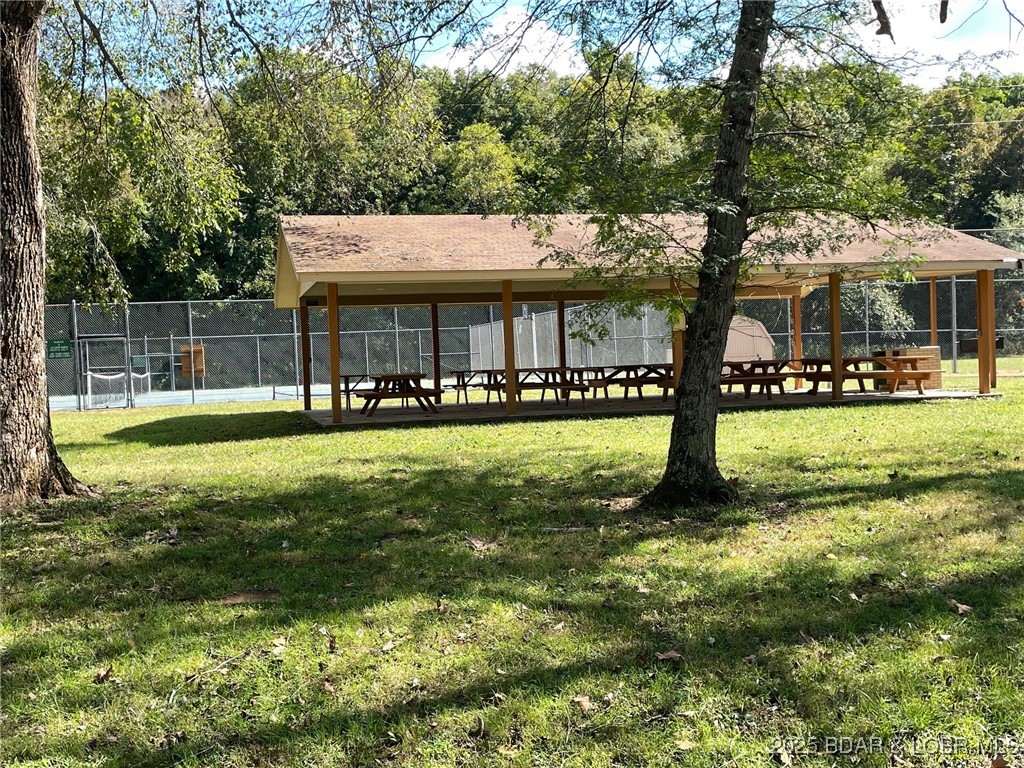 Covered picnic shelter with wooden tables beside a fenced tennis court, surrounded by trees.