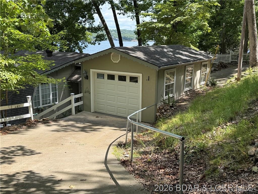 Garage and home on a wooded slope, with a concrete driveway and lake visible through the trees.