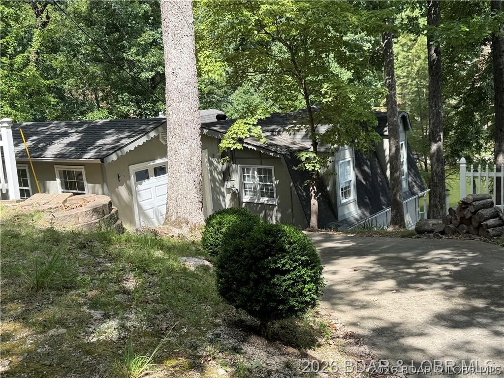 Gray house among mature trees, with a sloped driveway and stacked firewood.