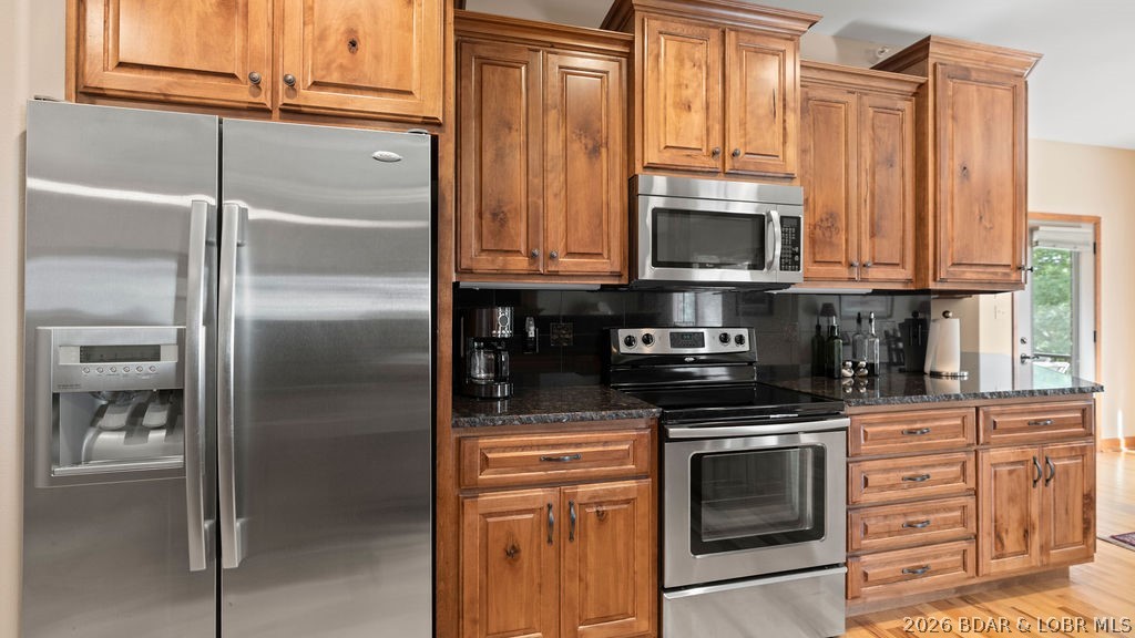 Kitchen with warm wood cabinets, dark countertops, stainless-steel appliances, and a window letting in daylight.
