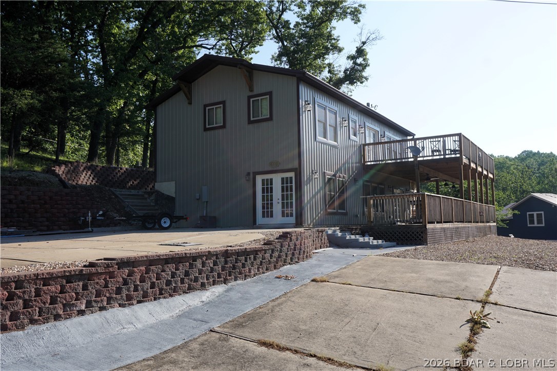 Gray two-story home with a large raised deck, retaining walls, and paved driveway.