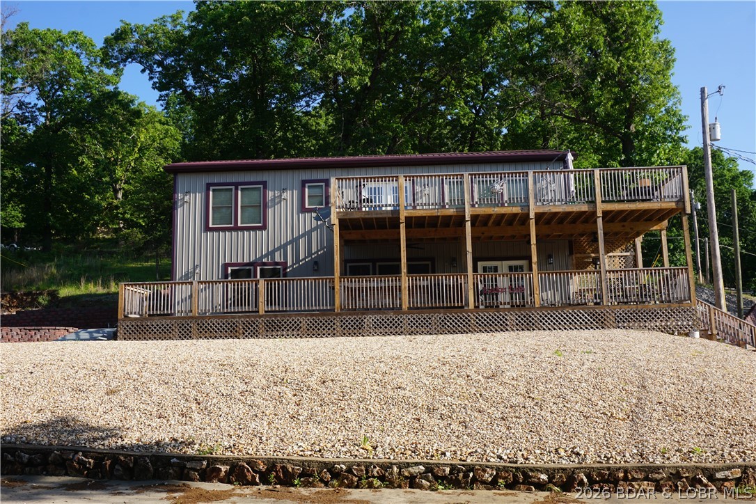 Gray two-story house with burgundy trim, expansive two-level deck, and gravel yard beneath leafy trees.