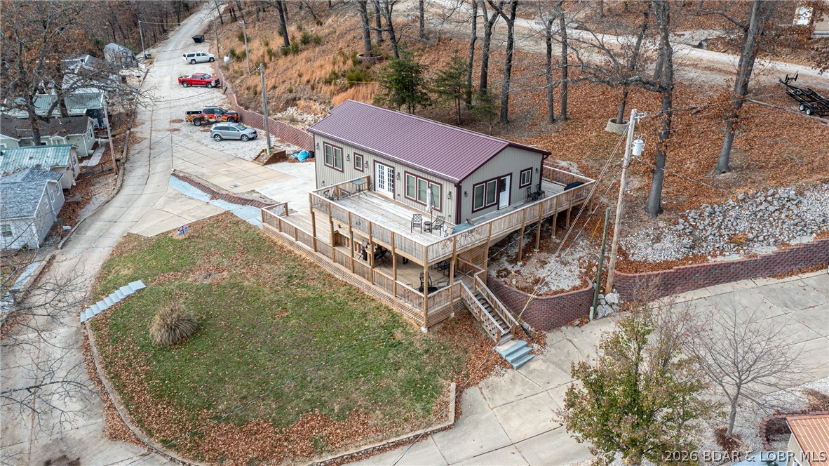 Aerial view of a single-story home with a raised wraparound deck beside a curved road.