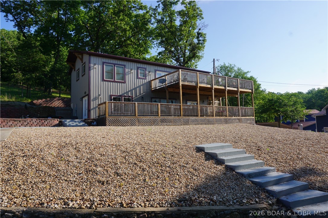 Two-story gray home with burgundy trim, a large wooden deck, and concrete steps across a gravel slope.
