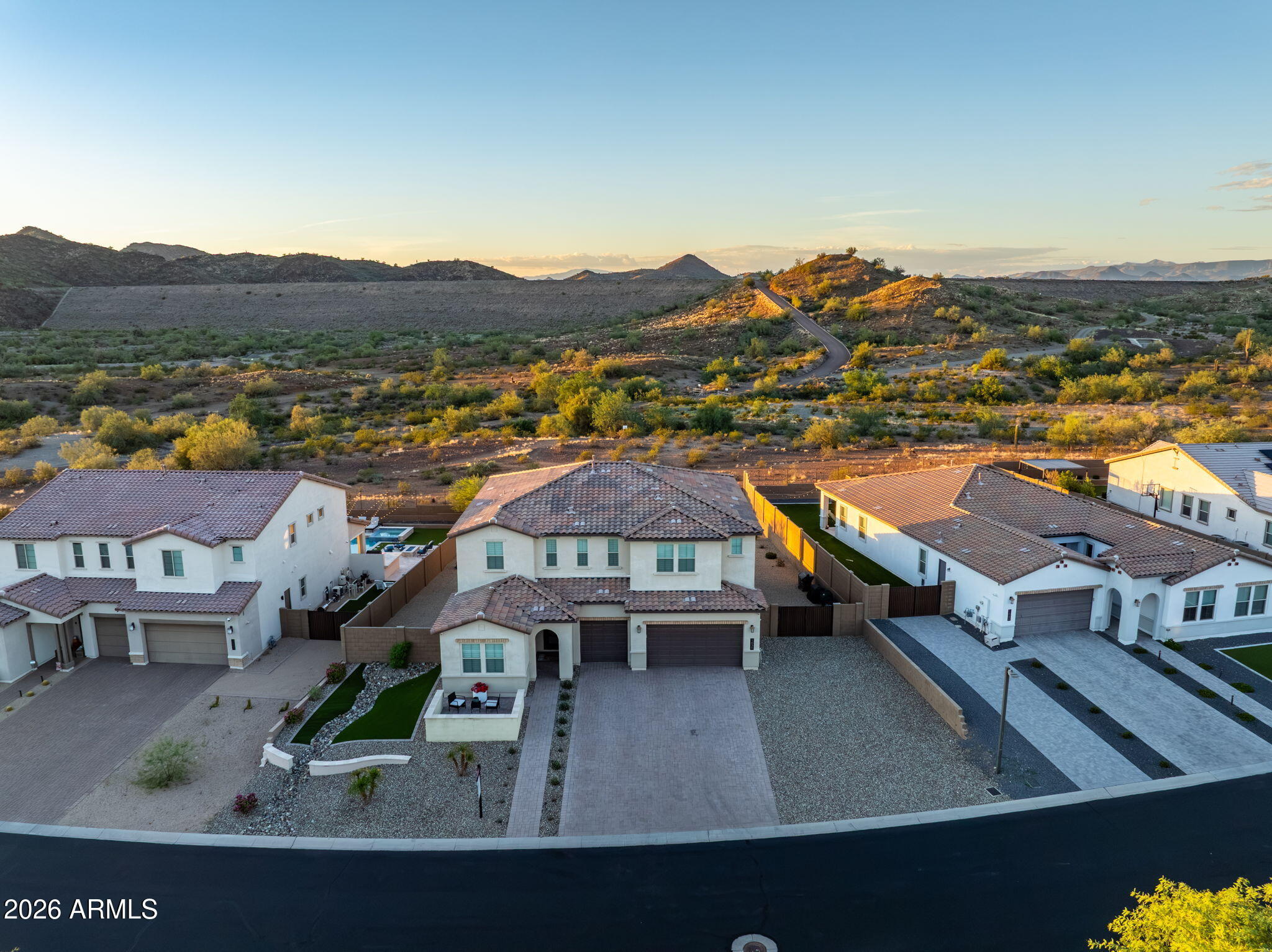 Aerial view of a stucco home with a paved driveway, neighboring homes, and desert mountains at golden hour.