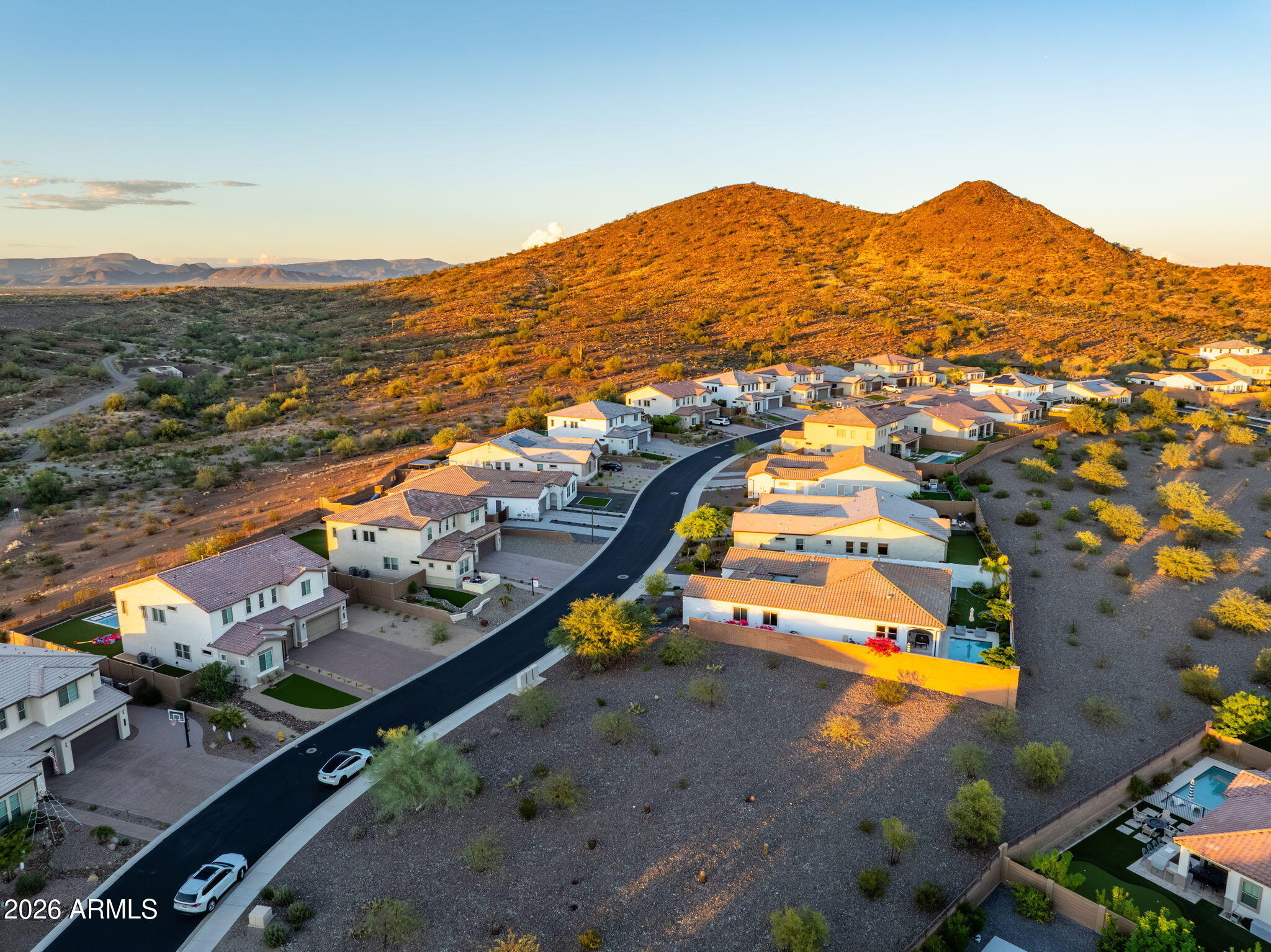 Aerial neighborhood view with a curving street, desert lots, and sunlit mountains behind the homes.